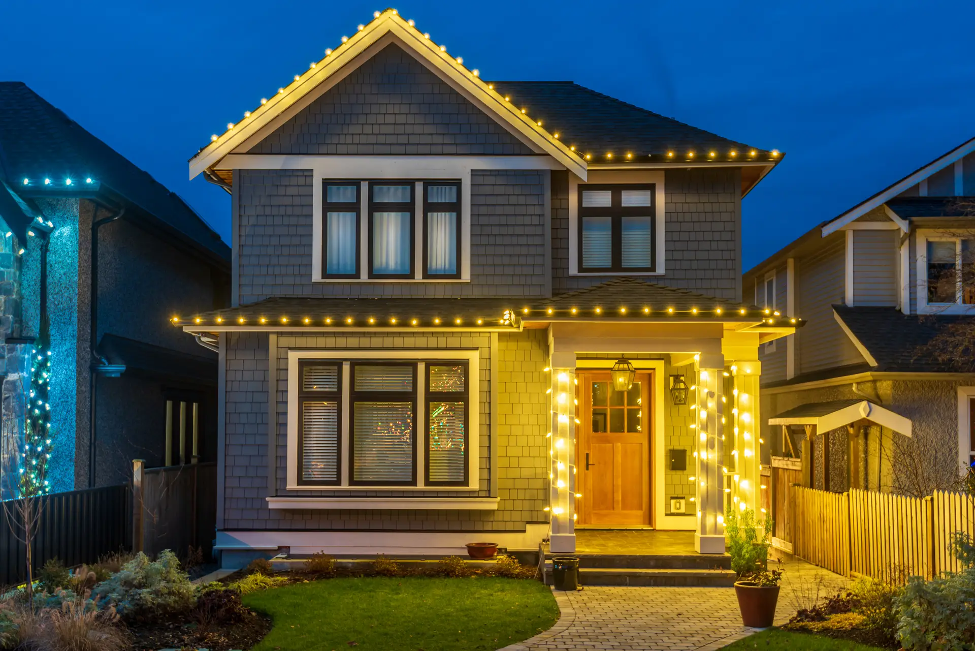 Two-storey home at blue hour with warm white lights on the roofline, porch and railings